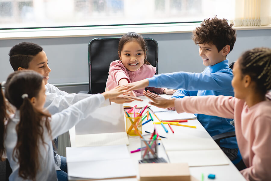 a group of smiling school kids sitting at a table in a classroom with their hands together in the center.