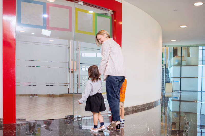 a mother and her young daughter walking through glass doors to enter a daycare center