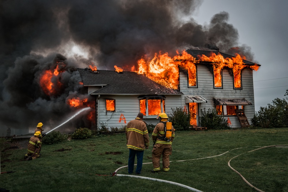 A suburban/rural house is seen engulfed in flames, as fire rages inside it, coming out the windows and creating dense smoke that spews above the home. Invest in residential fire safety protection to keep your property and your family safe.