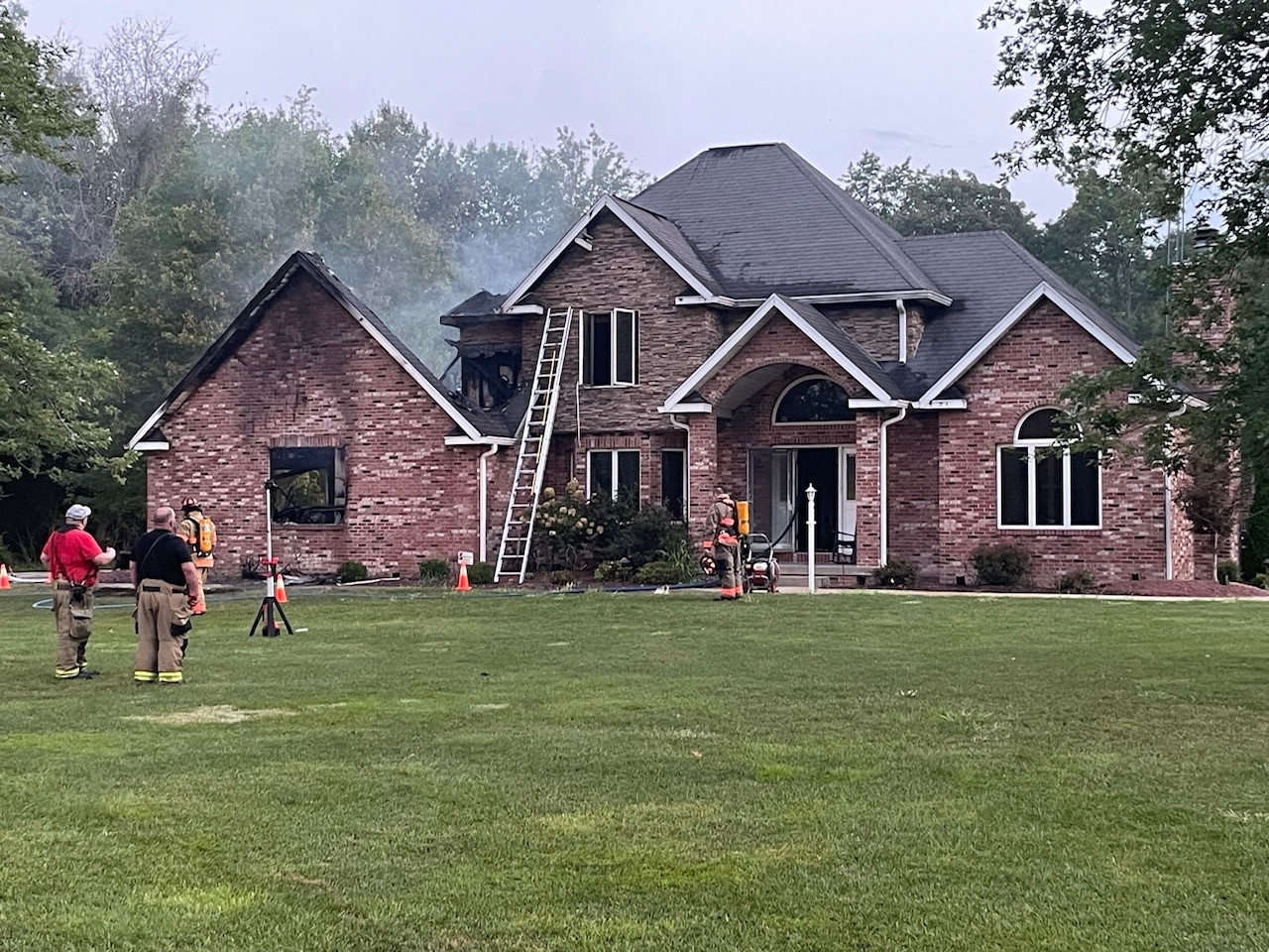 A house is seen after a fire, with a burnt roof and brick facade. Firefighters are seen working as smoke billows above the house.