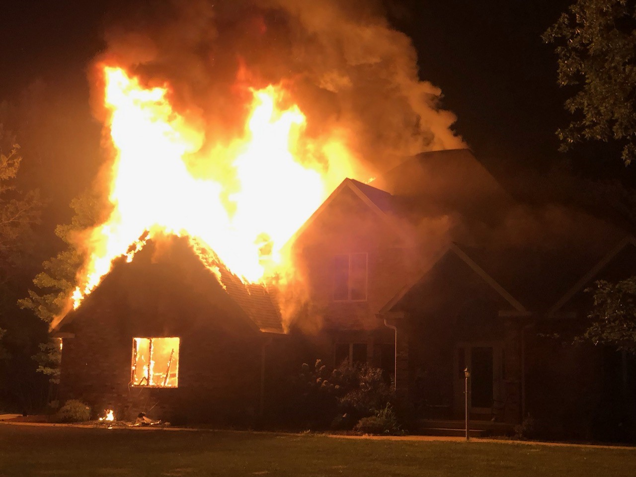 A house is seen engulfed in flames at nighttime. Orange flames can be seen inside the window and covering the roof.